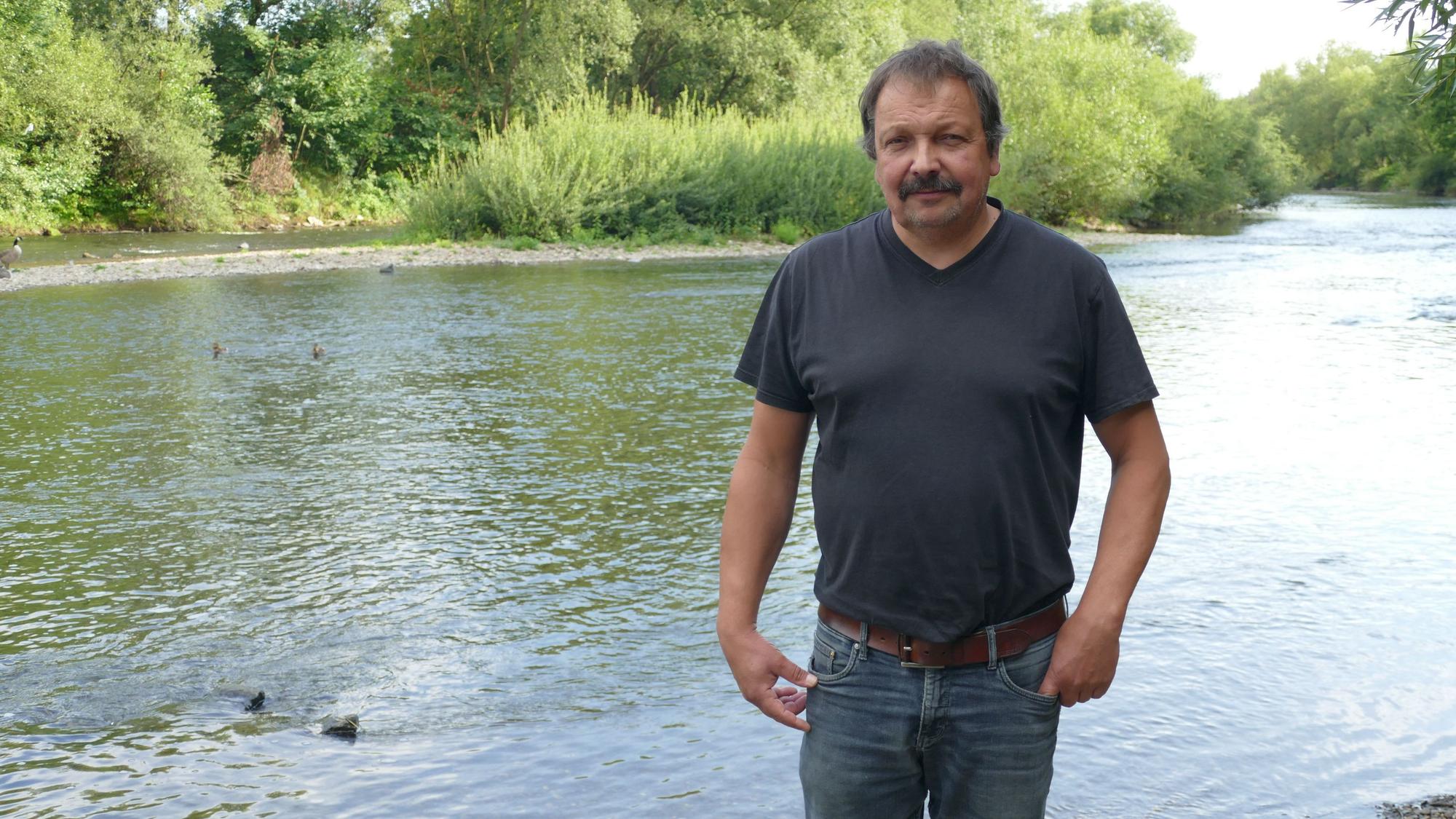 Ein Mann in T-Shirt und Jeans, im Hintergrund ist ein Fluss zu sehen.