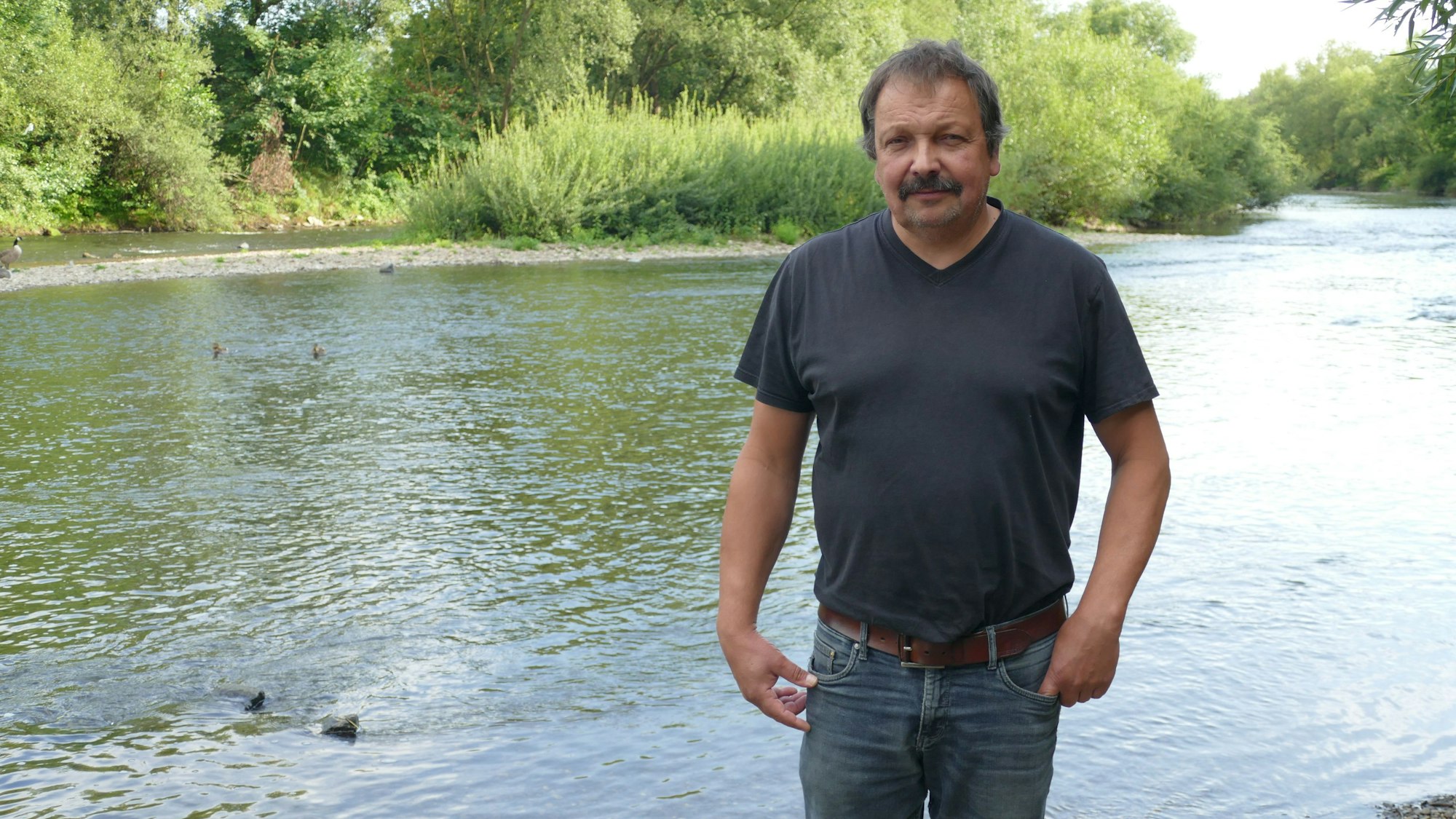 Ein Mann in T-Shirt und Jeans, im Hintergrund ist ein Fluss zu sehen.