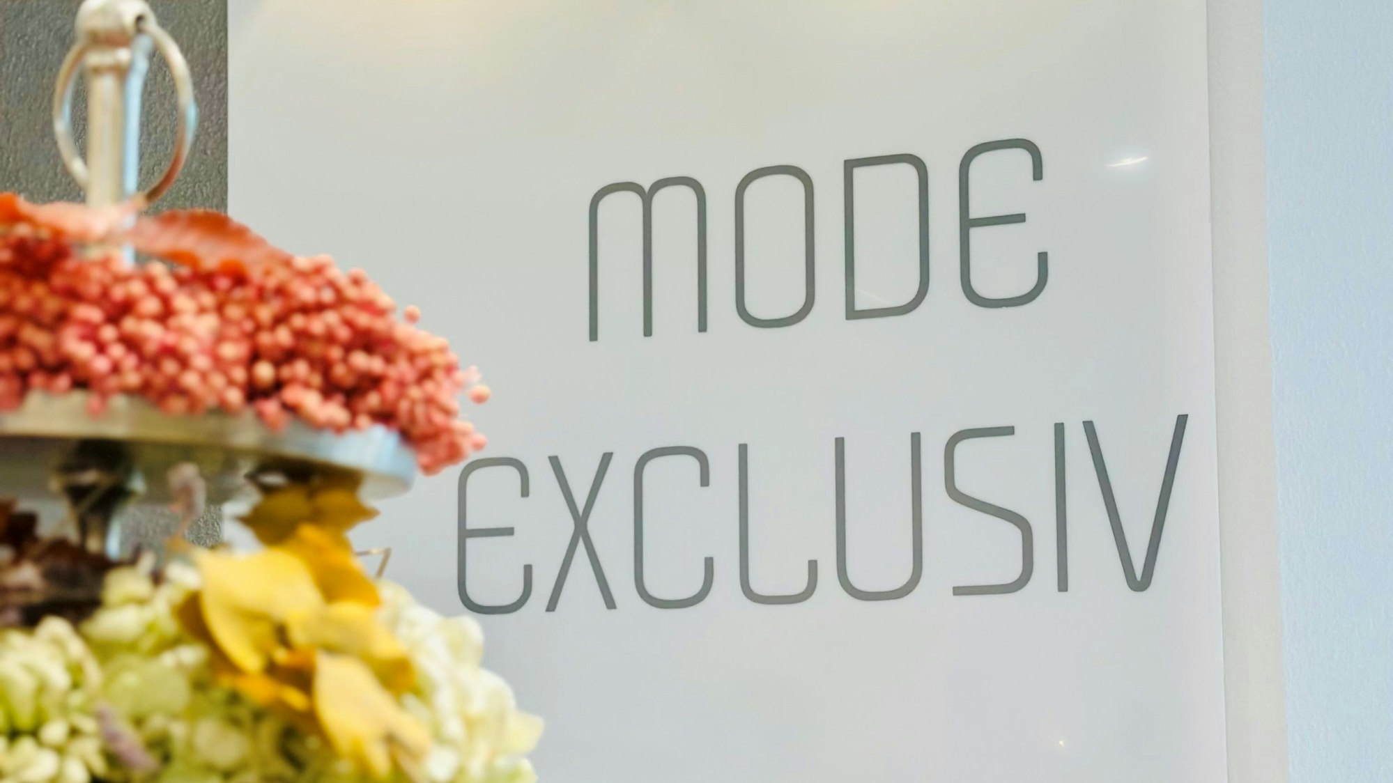 Zu sehen ist ein Schild mit dem Aufdruck „Mode Exclusiv“, davor stehen Blumen.