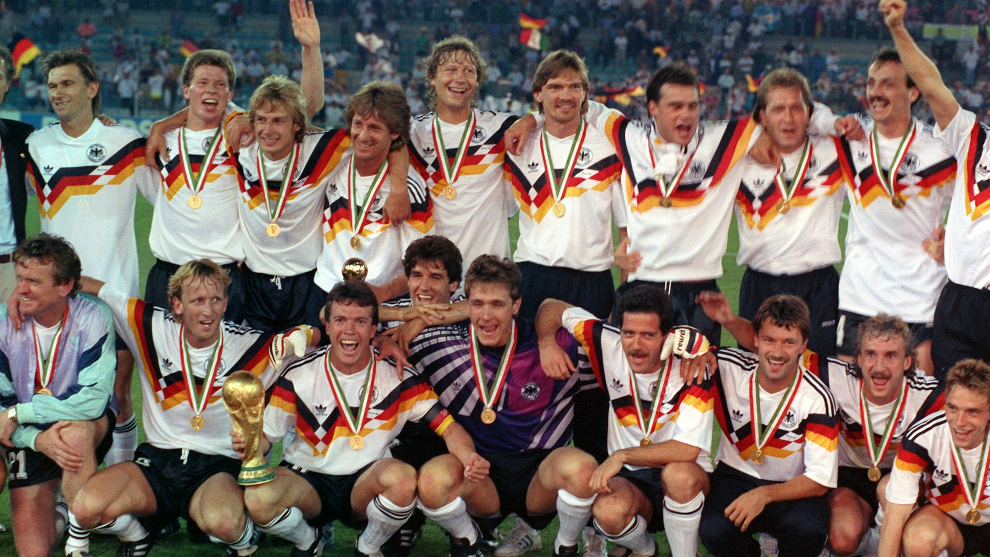 ARCHIV - 08.07.1990, Italien, Rom: Das Archivbild zeigt die deutsche Nationalmannschaft, die sich nach dem 1:0-Finalsieg gegen Argentinien bei der Fußball-Weltmeisterschaft im Olympiastadion von Rom zum Mannschaftsfoto aufstellt. Stehend (v.l.) Klaus Augenthaler, Stefan Reuter, Jürgen Klinsmann, Frank Mill, Guido Buchwald, Paul Steiner, Thomas Berthold, Andreas Köpke und Jürgen Kohler. Hockend (v.l.) Torwart-Trainer Sepp Maier, Andreas Brehme, Lothar Matthäus (mit dem WM-Pokal), Karlheinz Riedle, Bodo Illgner, Uwe Bein, Günter Hermann, Rudi Völler und Thomas Häßler. Deutschland wird nach 1954 und 1974 zum dritten Mal Weltmeister. Foto: picture-alliance / dpa +++ dpa-Bildfunk +++