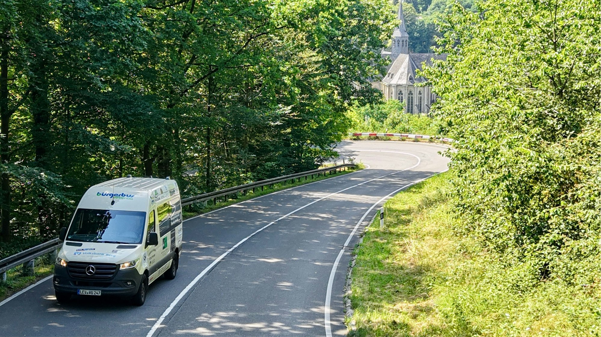 Ein kleiner Sprinter-Bus fährt durch die Serpentinen. Im Hintergrund sieht man den Altenberger Dom.