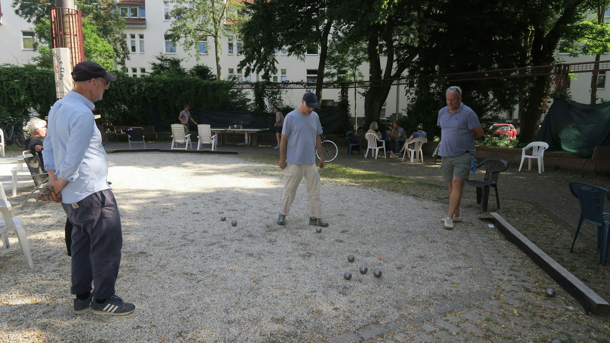 Drei Männer spielen Boule, im Hintergrund sitzen Menschen auf Plastikstühlen.