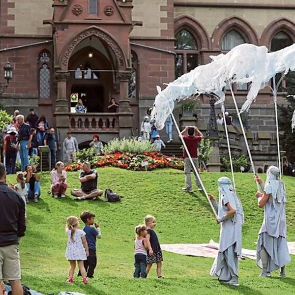 Drei verkleidete Gestalten auf einer Wiese vor einer Burg, einige Kinder schauen zu.
