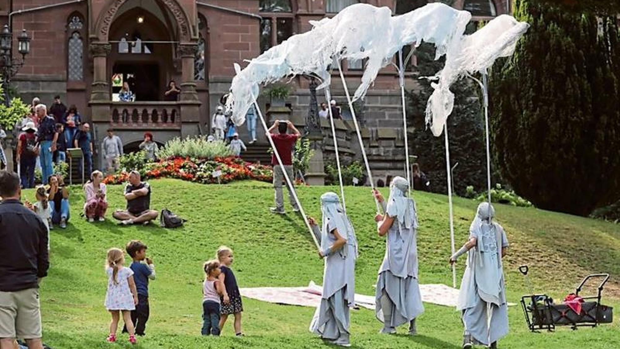 Drei verkleidete Gestalten auf einer Wiese vor einer Burg, einige Kinder schauen zu.