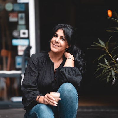 Eine Frau mit dunklem Haar im Pferdeschwanz sitzt auf den Stufen vor einer geöffneten Café-Tür.