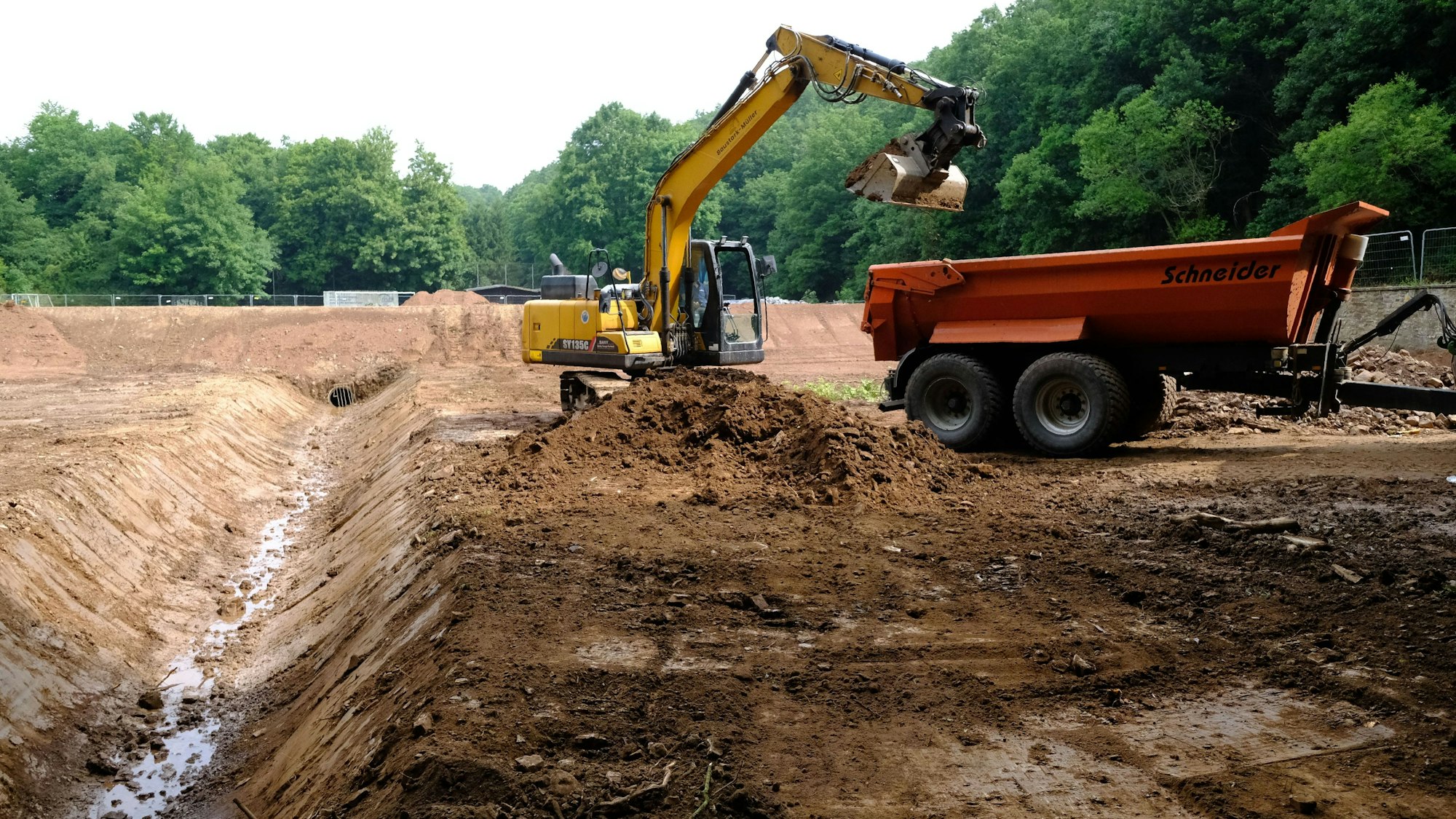 Ein Kran ist im Begriff, etwas auf einen Anhänger zuladen. Der Untergrund ist erdig und ausgetrocknet. Im Hintergrund stehen Bäume.