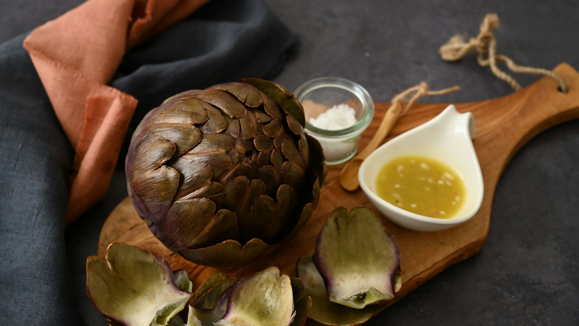 Artischocken haben jetzt Saison. Eine zubereitete Artischocke liegt auf einem Brettchen, daneben eine Schale Vinaigrette.