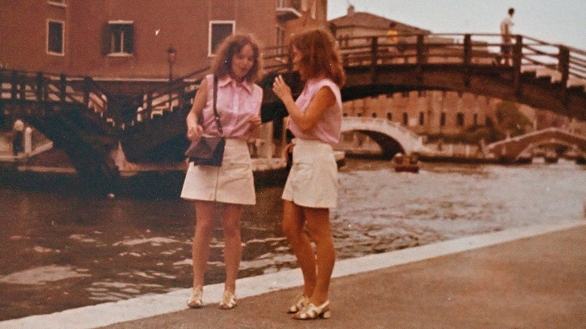 Die Zwillinge stehen vor einer Brücke und tragen beige Röcke und rosa Blusen.
