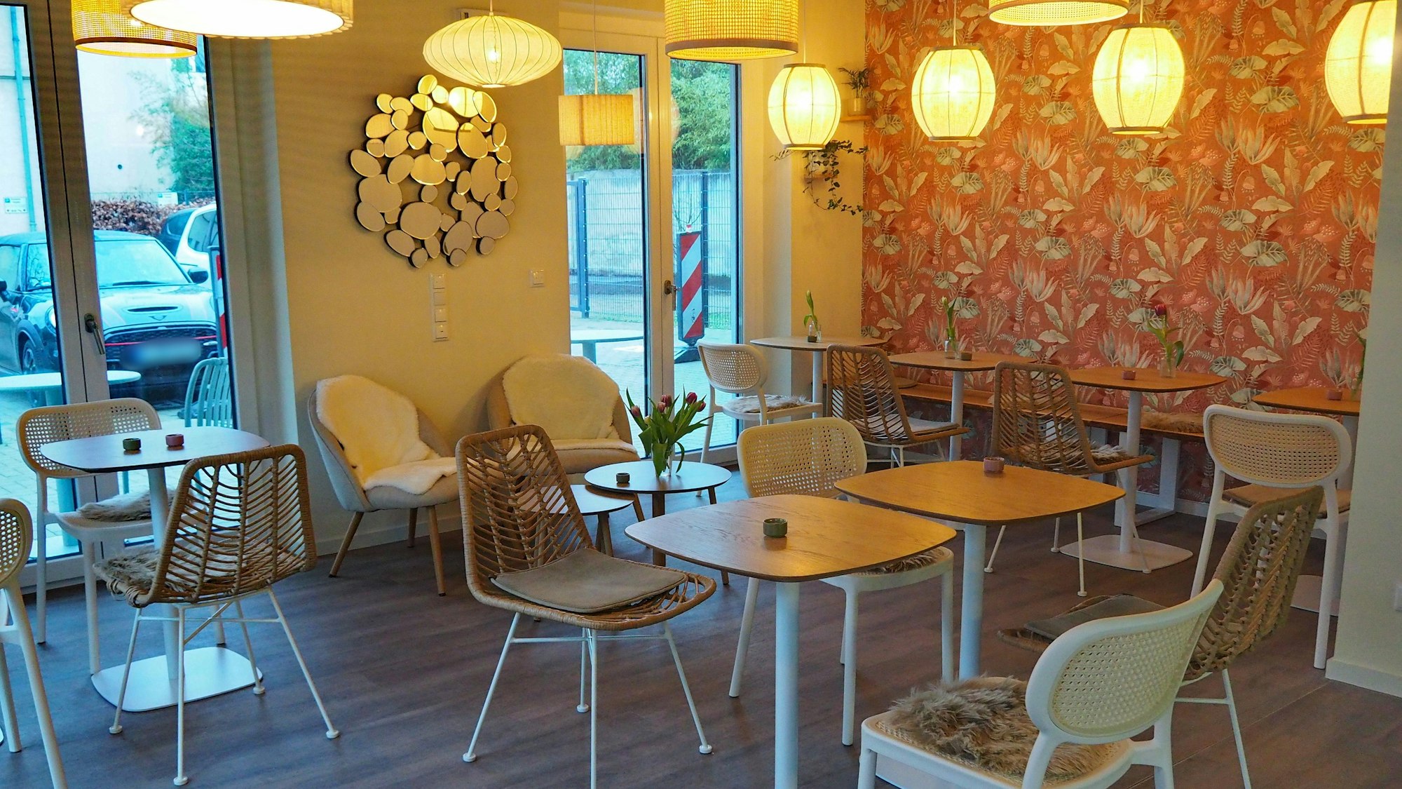 Tische und Stühle stehen in einem Café mit Blümchentapete an der einen und einem Spiegelkunstwerk an einer anderen Wand.