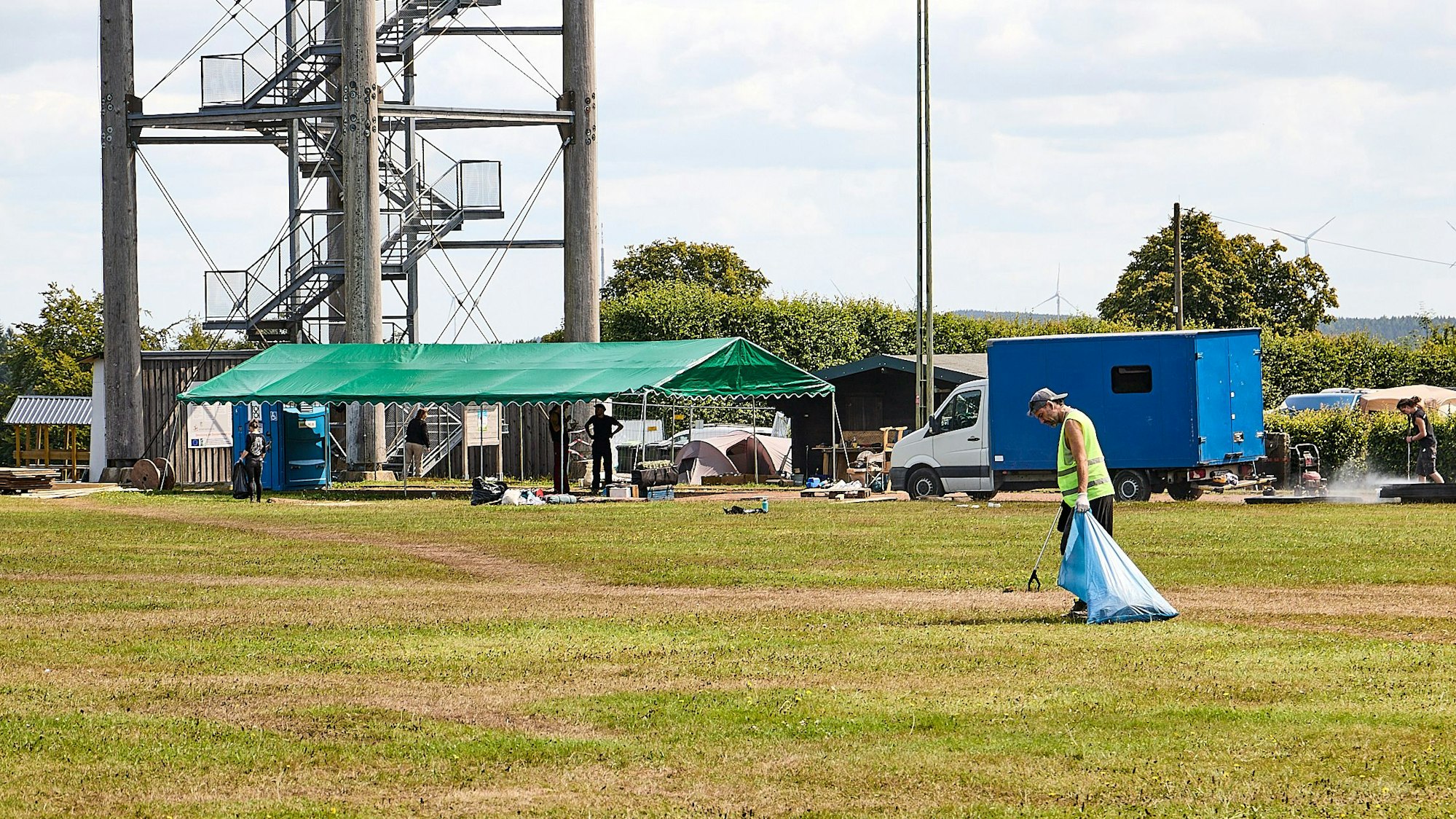 Mit Zange und Müllsack ist Nils auf dem Sportplatz unterwegs. Im Hintergrund sind der Aussichtsturm und das Festival-Gelände zu sehen.