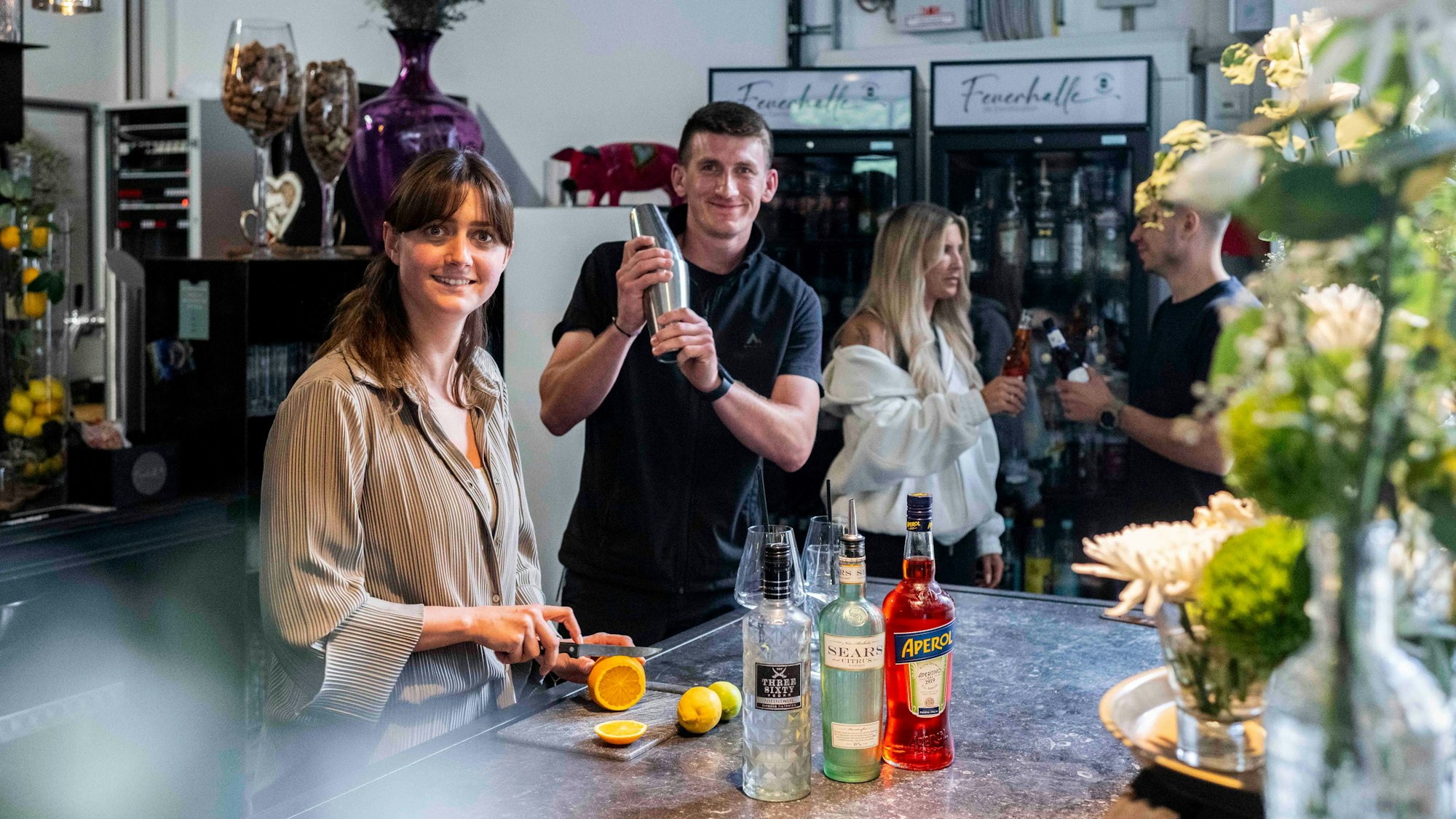 Linda-Marie Hannes und Max Hannewald stehen hinter einem Tresen. Sie schneidet Orangen und er mixt etwas in einem Shaker.