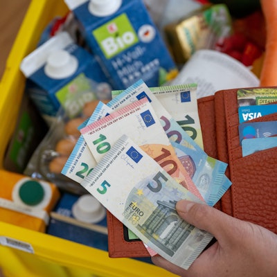 Eine Frau hält ein Portemonnaie mit Bargeld und Karten über einer Einkaufskiste mit Lebensmitteln.