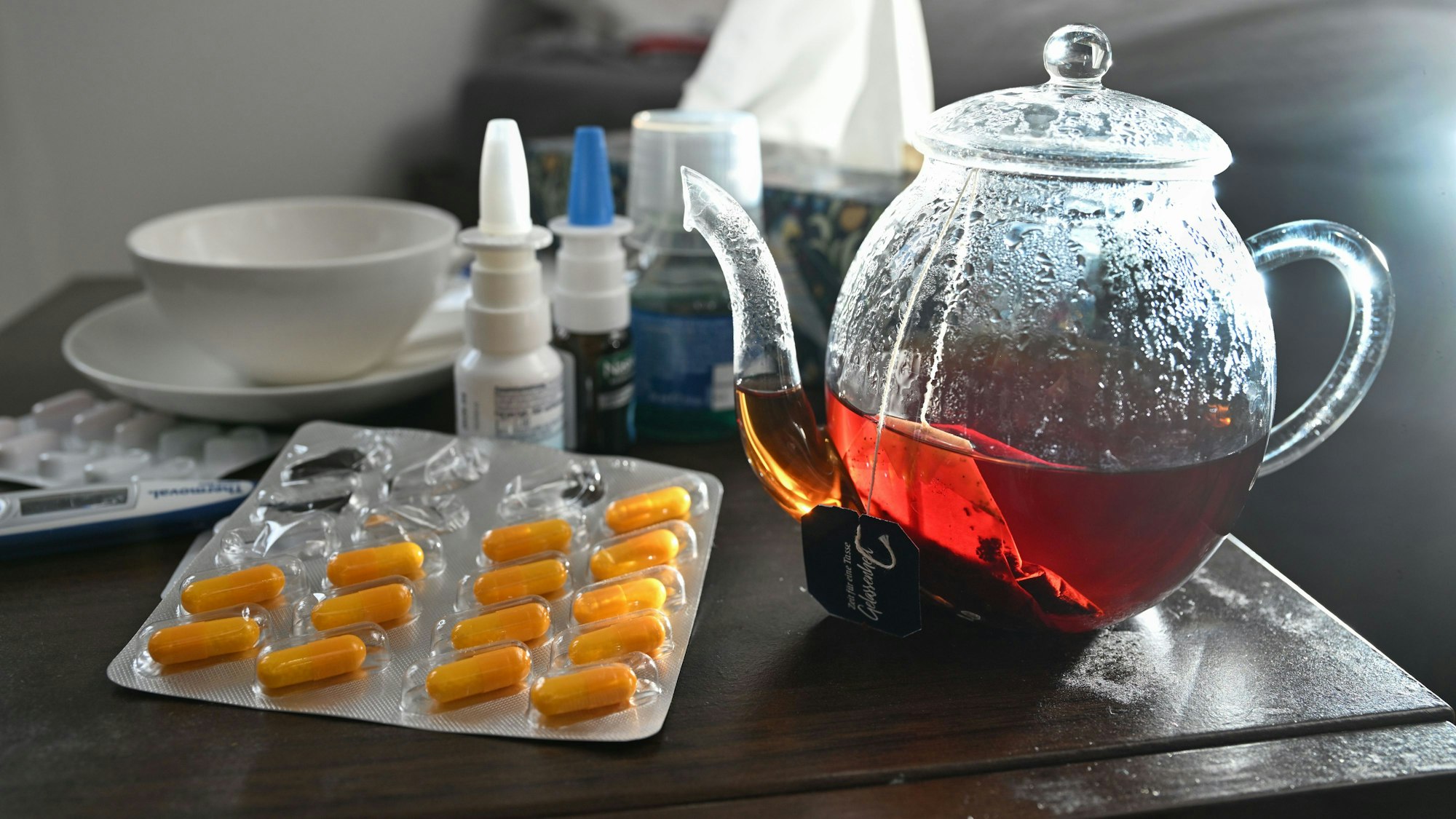 Medikamente und eine Kanne Tee zur Bekämpfung einer Erkältung sind auf einem Nachttisch aufgestellt.