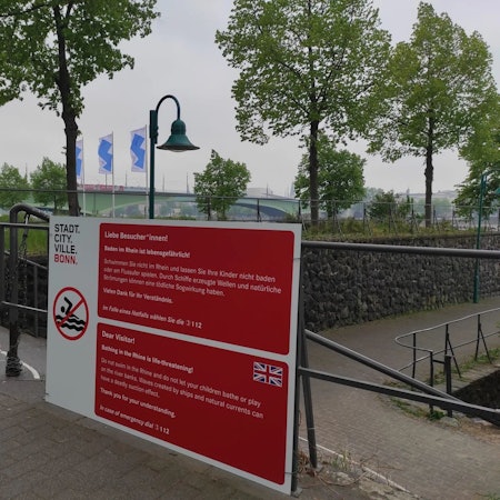 Ein Hinweisschild der Stadt Bonn in den Farben rot und weiß. Der Text lautet: „Liebe Besucher*innen! Baden im Rhein ist lebensgefährlich! Schwimmen Sie nicht im Rhein und lasse Sie Ihre Kinder nicht baden oder am Flussufer spielen Durch Schiffe erzeugte Wellen und natürliche Strömungen können einen tödliche Sogwirkung haben. Vielen Dank für Ihr Verständnis. Im Falle eines Notfalls wählen Sie die 112.“