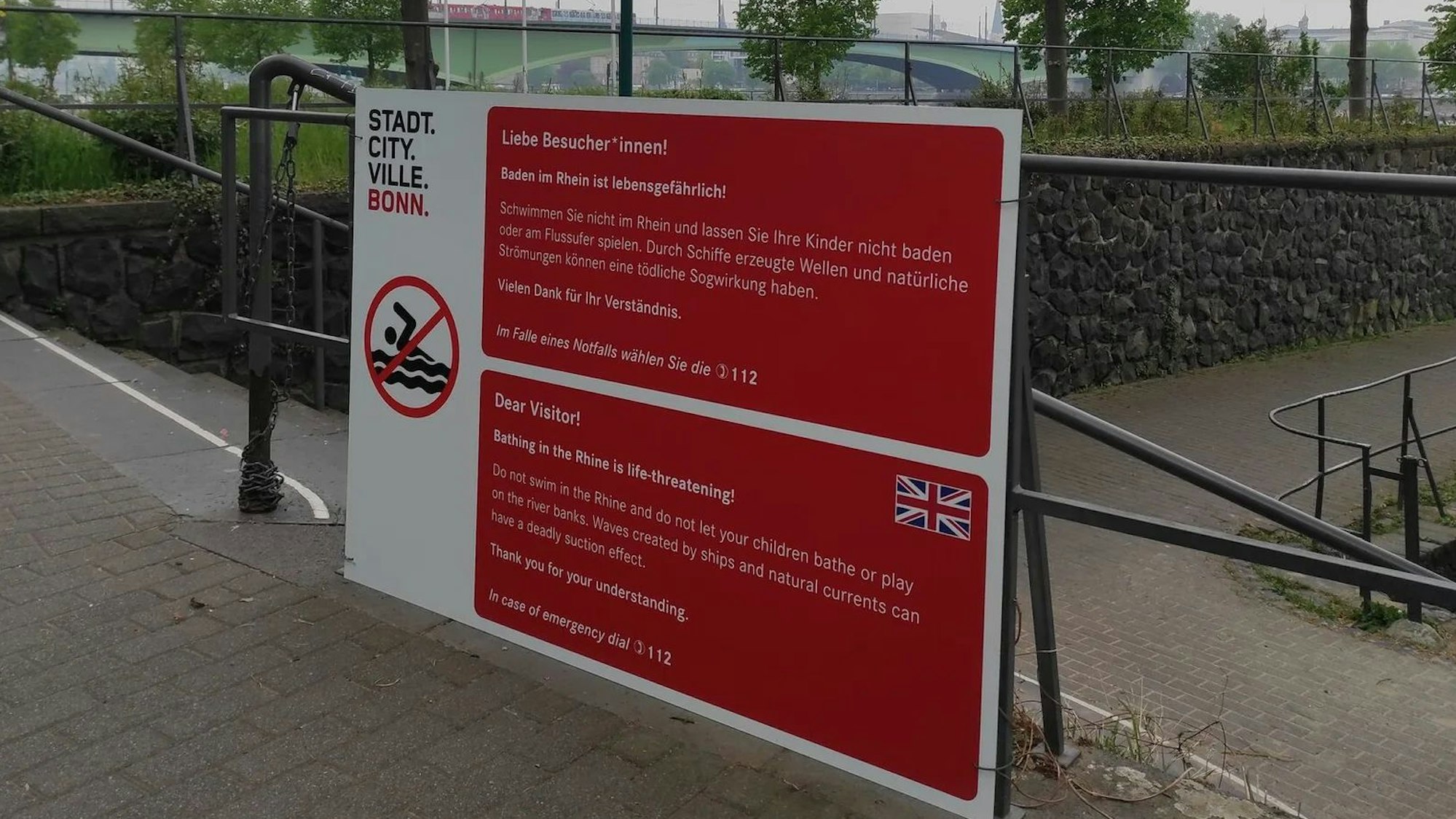 Ein Hinweisschild der Stadt Bonn in den Farben rot und weiß. Der Text lautet: „Liebe Besucher*innen! Baden im Rhein ist lebensgefährlich! Schwimmen Sie nicht im Rhein und lasse Sie Ihre Kinder nicht baden oder am Flussufer spielen Durch Schiffe erzeugte Wellen und natürliche Strömungen können einen tödliche Sogwirkung haben. Vielen Dank für Ihr Verständnis. Im Falle eines Notfalls wählen Sie die 112.“