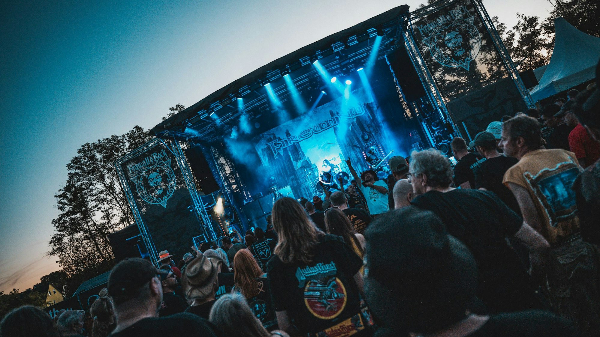 Publikum steht vor einer Bühne im Freien, die von verschiedenen Strahlern in ein blaues Licht getaucht wird.