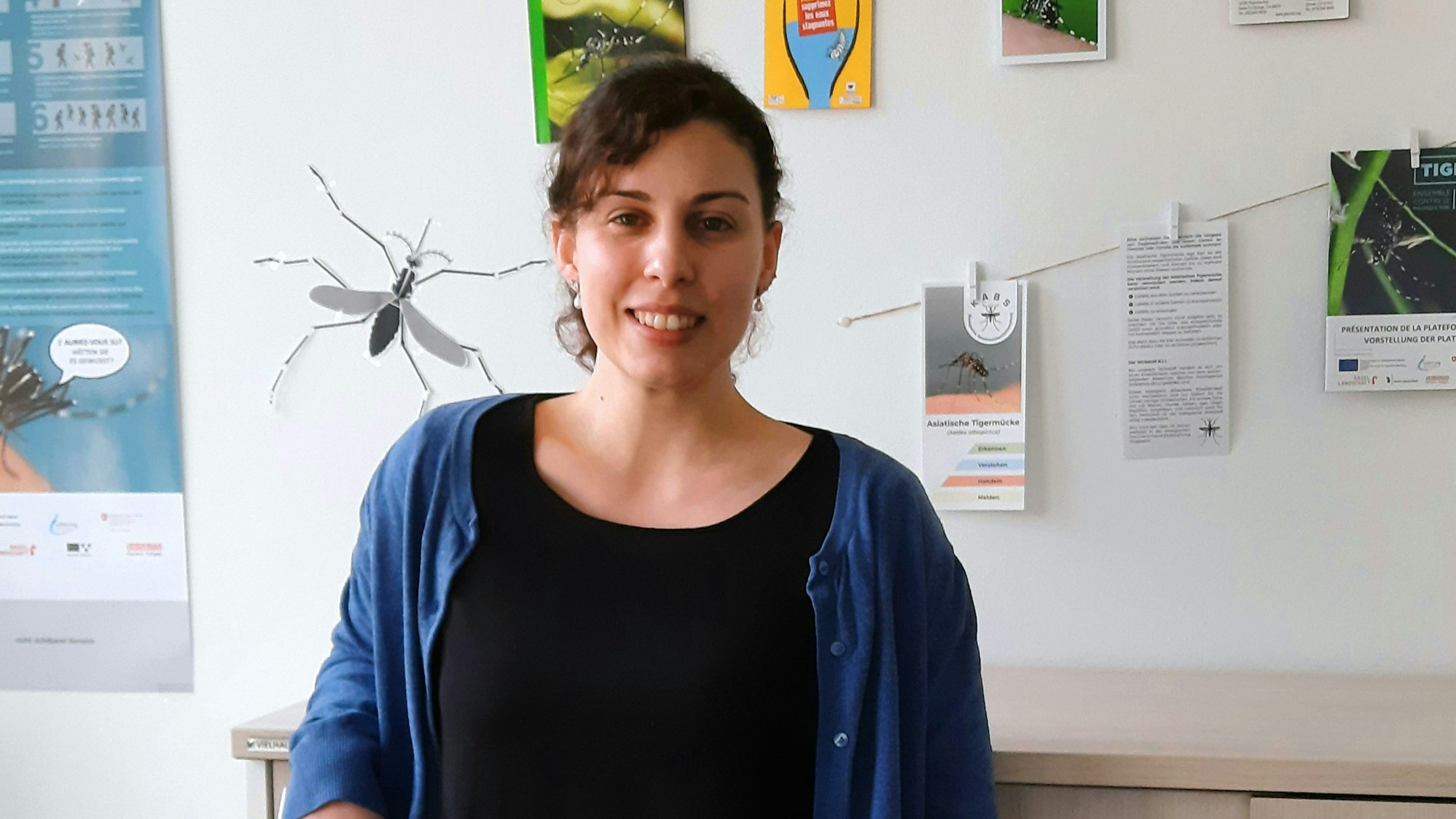 Frau Augsten ist in ihrem Büro vor einer mit Plakaten vollgehängten Wand zu sehen. Sie lächelt in die Kamera.