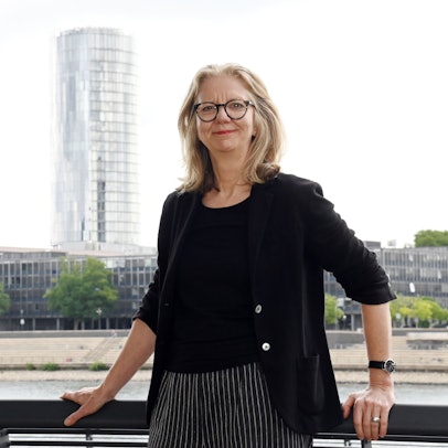 Architektin Dörte Gatermann steht am Pegel Köln, im Hintergrund ist das Köln-Triangle-Hochhaus zu sehen.