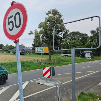 Zu sehen ist eine Straße und daneben ein Metallrahmen, in dem ein Ortseingangsschild fehlt, sowie ein provisorisch aufgestelltes Tempo-50-Schild.