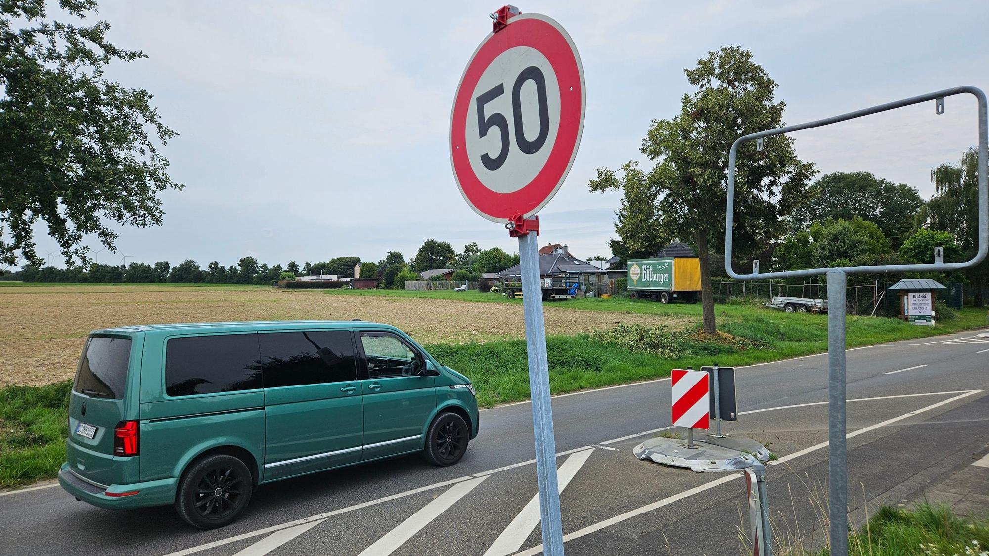 Zu sehen ist eine Straße und daneben ein Metallrahmen, in dem ein Ortseingangsschild fehlt, sowie ein provisorisch aufgestelltes Tempo-50-Schild.