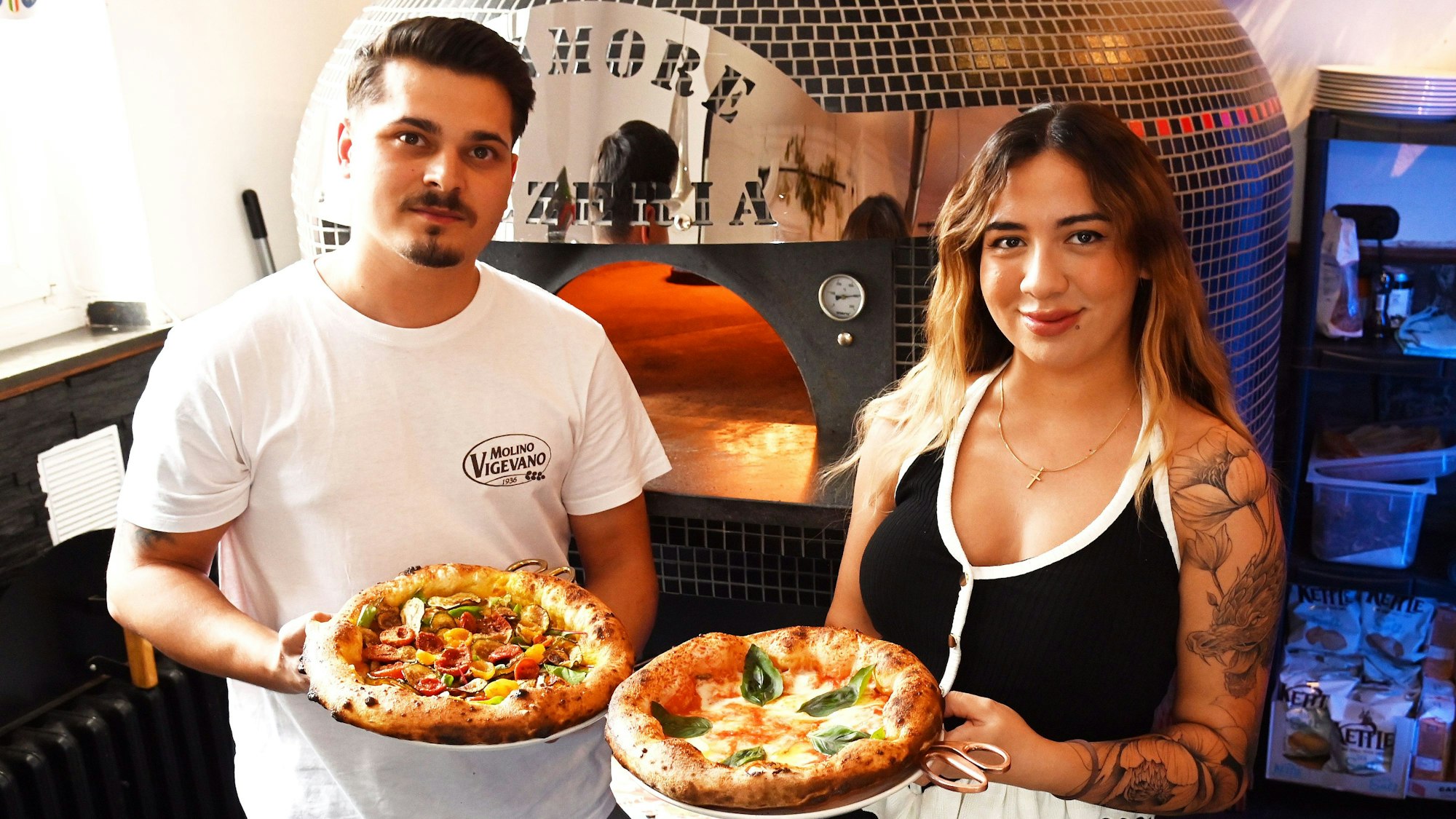 Die beiden Gastgeber sind vor dem Pizzaofen zu sehen. Beide zeigen eine frisch gebackene Pizza.