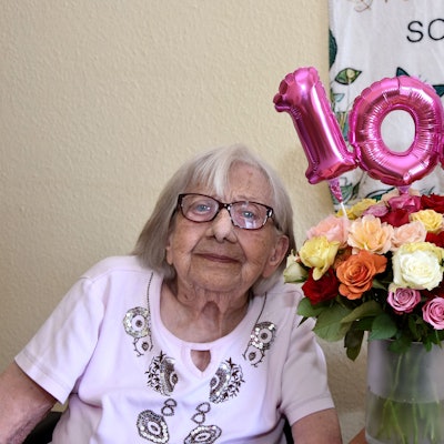 Neben der Seniorin steht eine Vase mit Blumen, darüber die Zahl 100 als Luftballon.