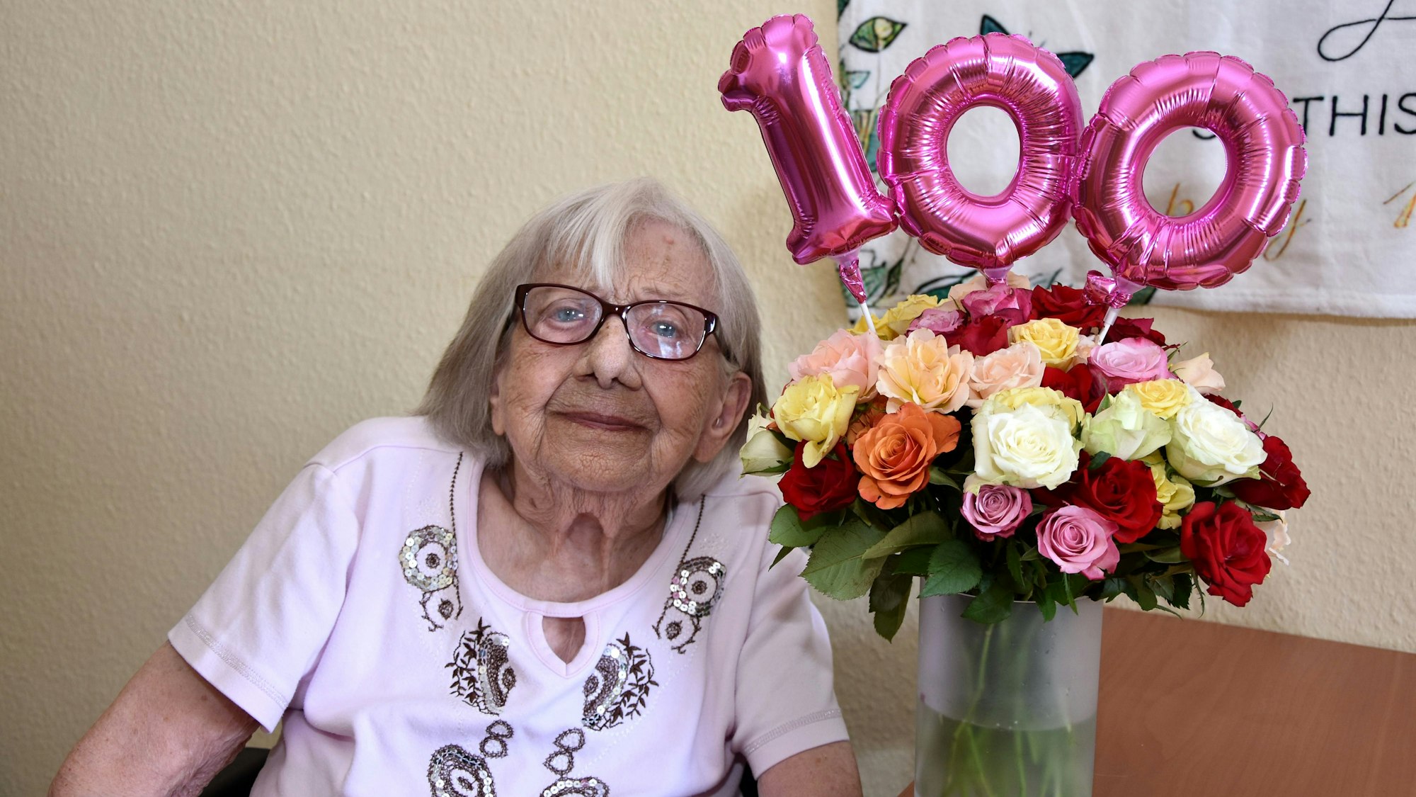 Neben der Seniorin steht eine Vase mit Blumen, darüber die Zahl 100 als Luftballon.