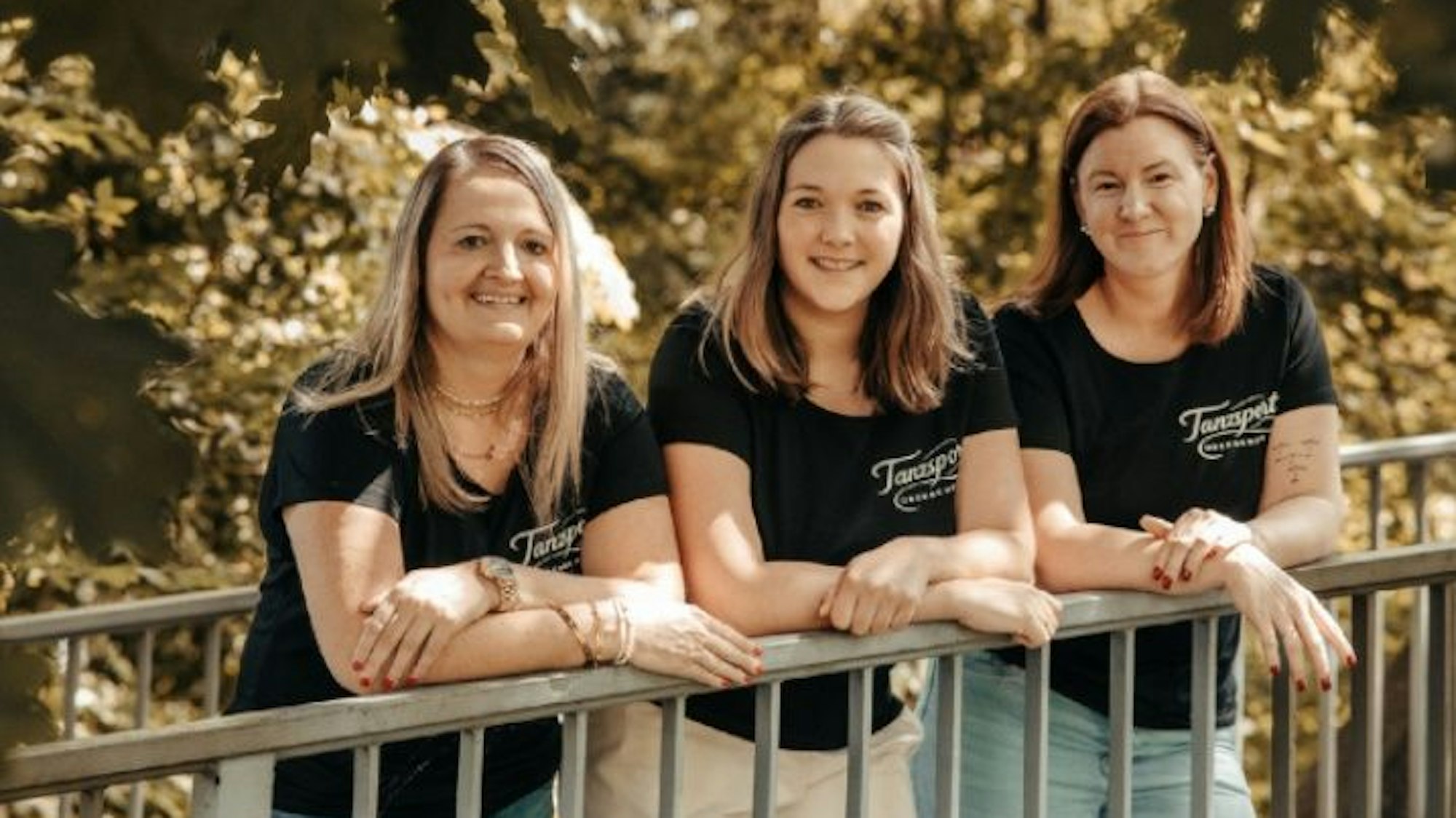 Drei Frauen lehnen lässig auf einem Geländer und tragen dabei das gleiche T-Shirt.