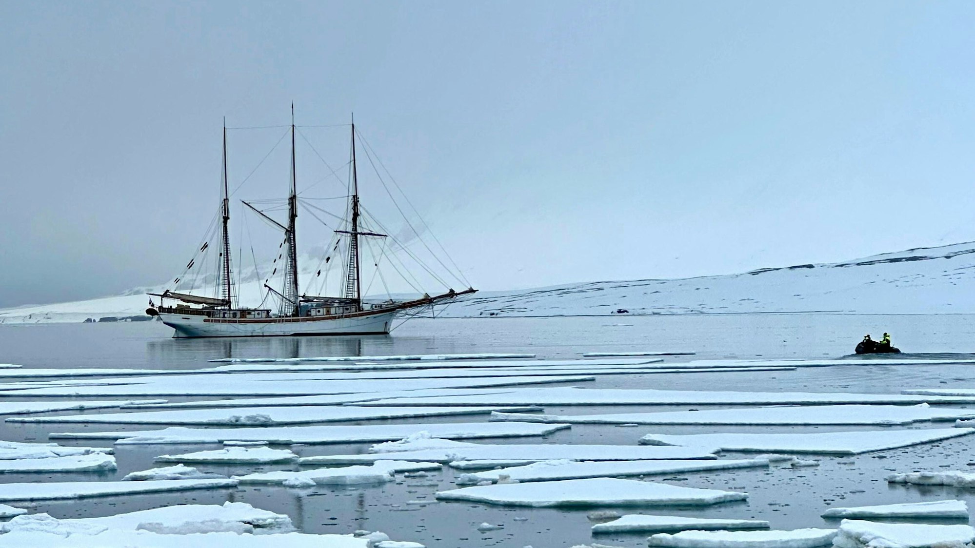 Ein dreimastiges Segelschiff liegt vor der Küste vor Anker. Im Vordergrund schwimmen Eisschollen auf dem Wasser.