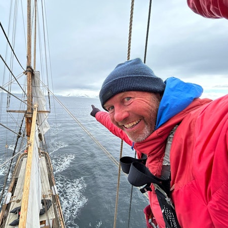André Hauschke trägt eine rote Daunenjacke und blaue Wollmütze und deutet an Bord eines Schiffes in Richtung Horizont.