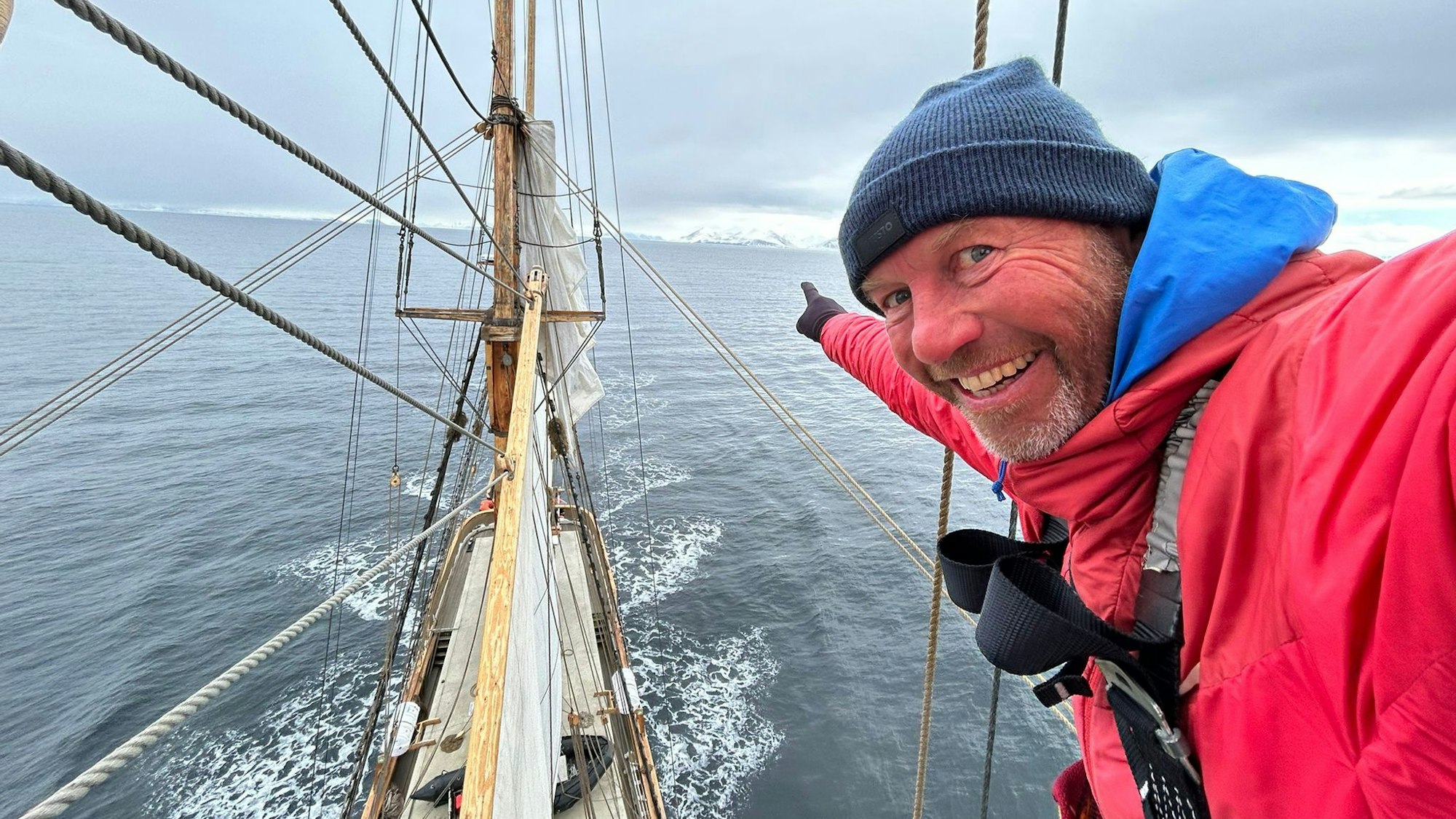 André Hauschke trägt eine rote Daunenjacke und blaue Wollmütze und deutet an Bord eines Schiffes in Richtung Horizont.
