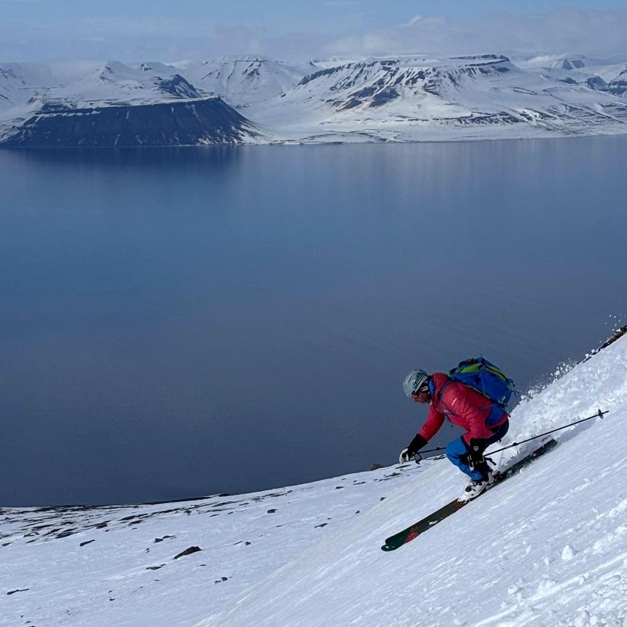 Ein Skifahrer in roter Daunenjacke bei der Abfahrt. Im Hintergrund das Meer und eine weitere schneebedeckte Insel.
