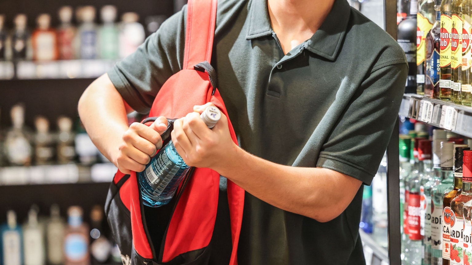 Ein Mann steckt in einem Supermarkt eine Flasche mit einem alkoholhaltigen Getränk in seinen Rucksack. (Symbolbild)