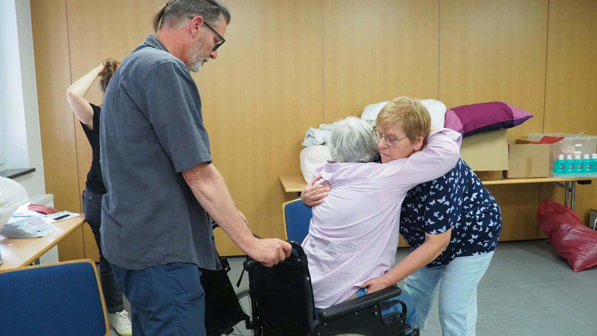 Christine fasst Marita um Schulter und Hüfte und lässt sie auf den Sitz des Rollstuhls gleiten. Georg sichert den Stuhl an den Griffen.