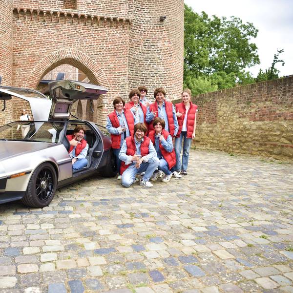 Sieben Mitglieder der Karnevalisten stehen mit Perücken neben einem silbernen Rennwagen mit nach oben geöffneten Flügeltüren. Ein Fahrer sitzt am Steuer.