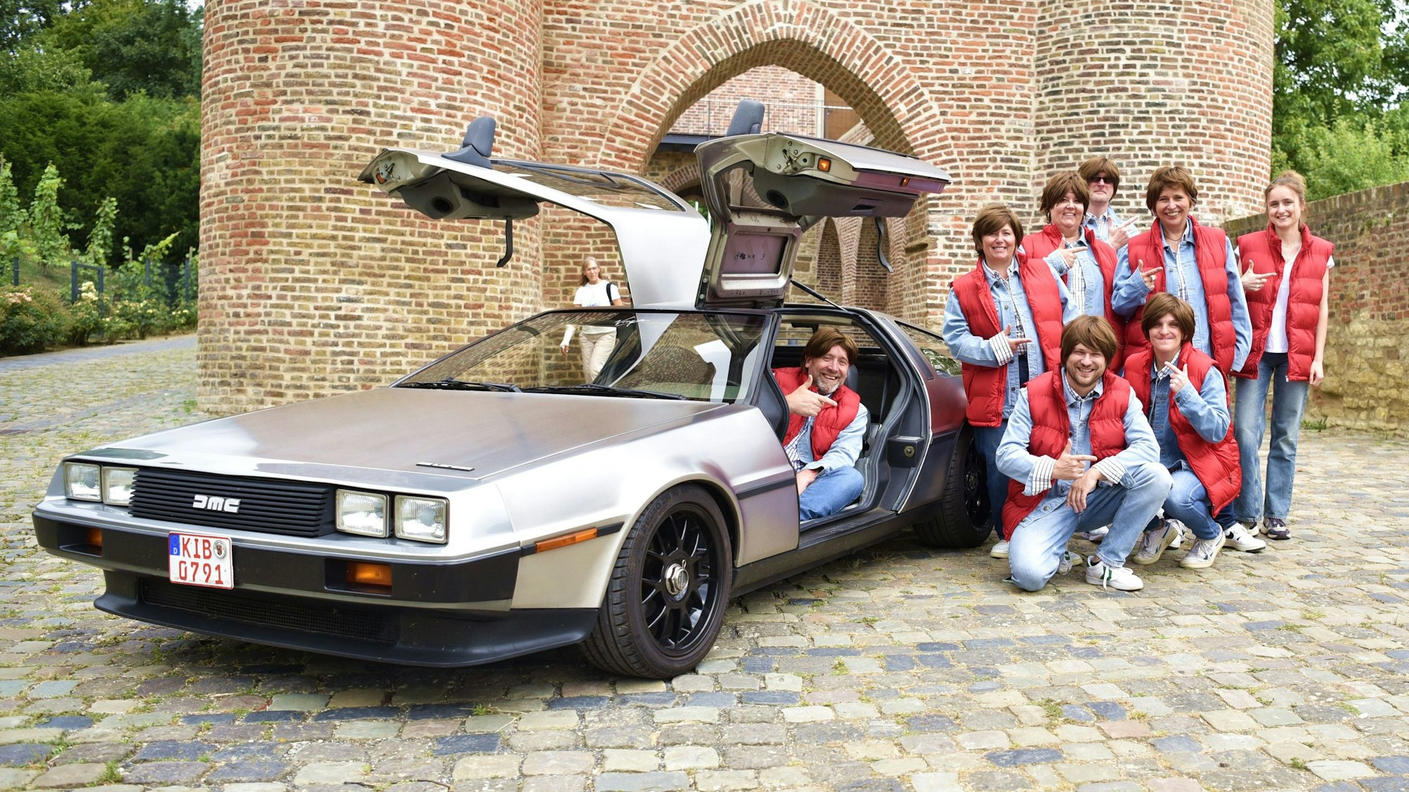 Sieben Mitglieder der Karnevalisten stehen mit Perücken neben einem silbernen Rennwagen mit nach oben geöffneten Flügeltüren. Ein Fahrer sitzt am Steuer.