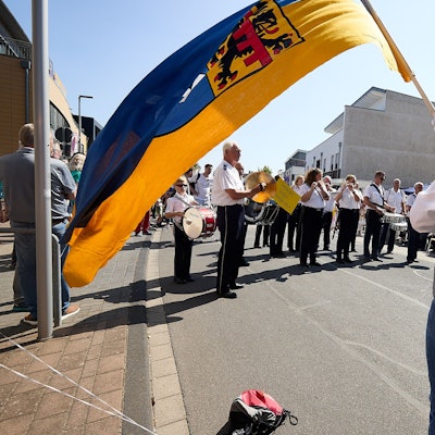 Blick unter einer großen Stadtfahne, die von einem Mann mit Zylinder gehalten wird, hindurch auf die Musikkapelle.