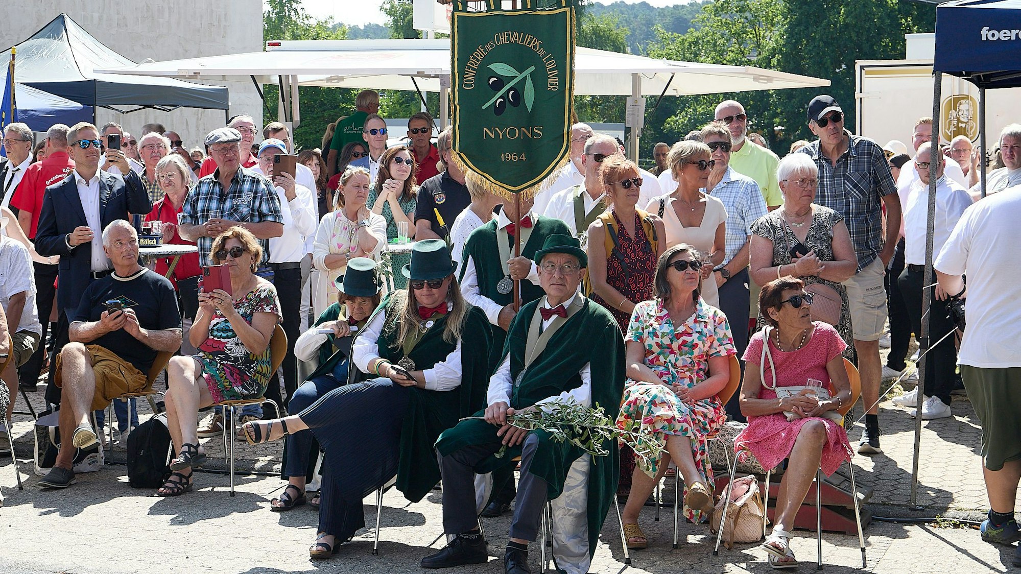 Drei neue Ritter sitzen mit Zylindern und dunklen Umhängen auf Stühlen unter einer Fahne. Dahinter stehen Festbesucher.