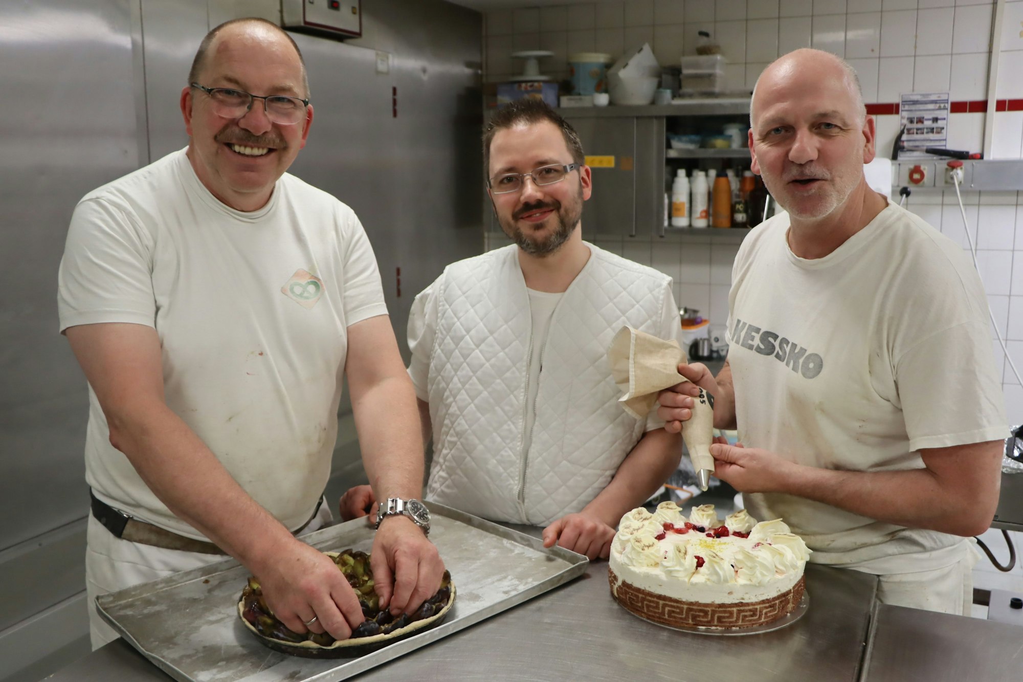Alle drei Männer tragen helle Kleidung. Links macht Alexander Kayer einen Obstkuchen fertig, rechts im Bild sprüht Stephan Gärtner Sahne auf eine Torte.