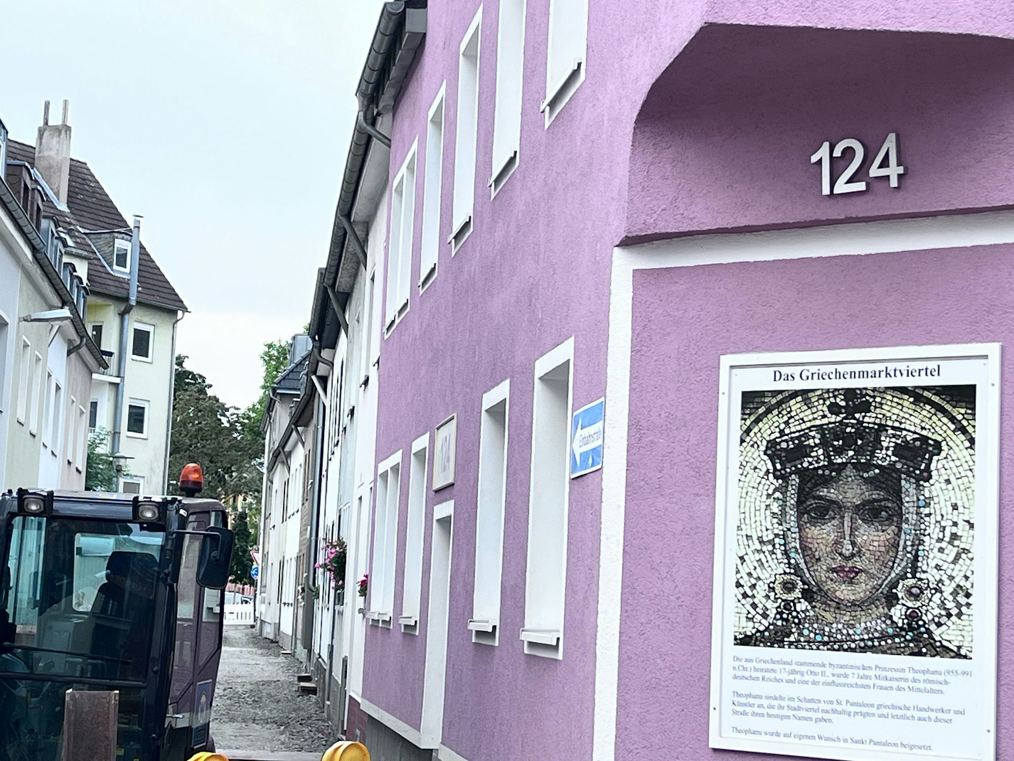 Eine kleine Gasse, in denen gerade Bauarbeiten stattfinden, ist zu sehen. Im Vordergrund hängt eine Erklärtafel an einem Haus mit lilafarbener Fassade zum Namen Griechenmarktviertel.