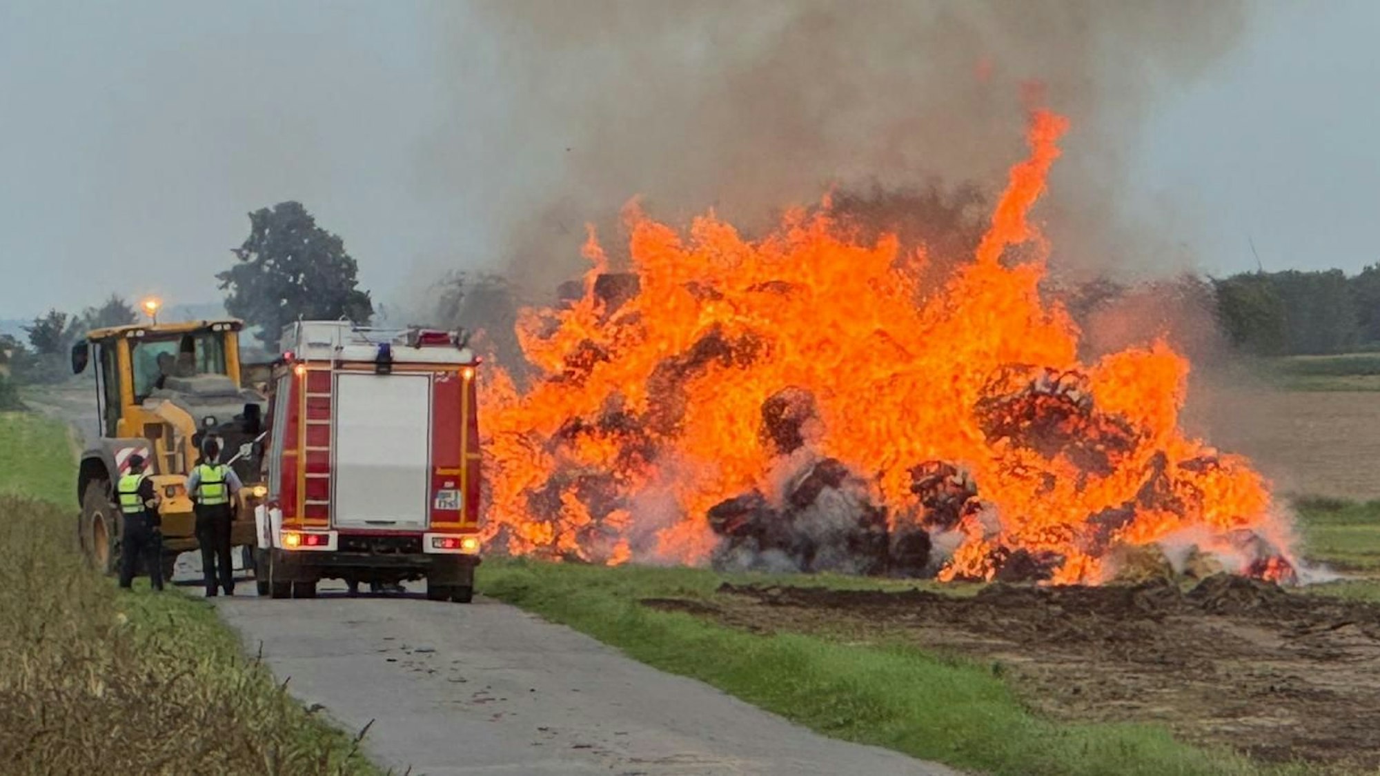 Ein Haufen Strohballen in Flammen. Daneben ein Traktor, ein Feuerwehrfahrzeug und Einsatzkräfte.