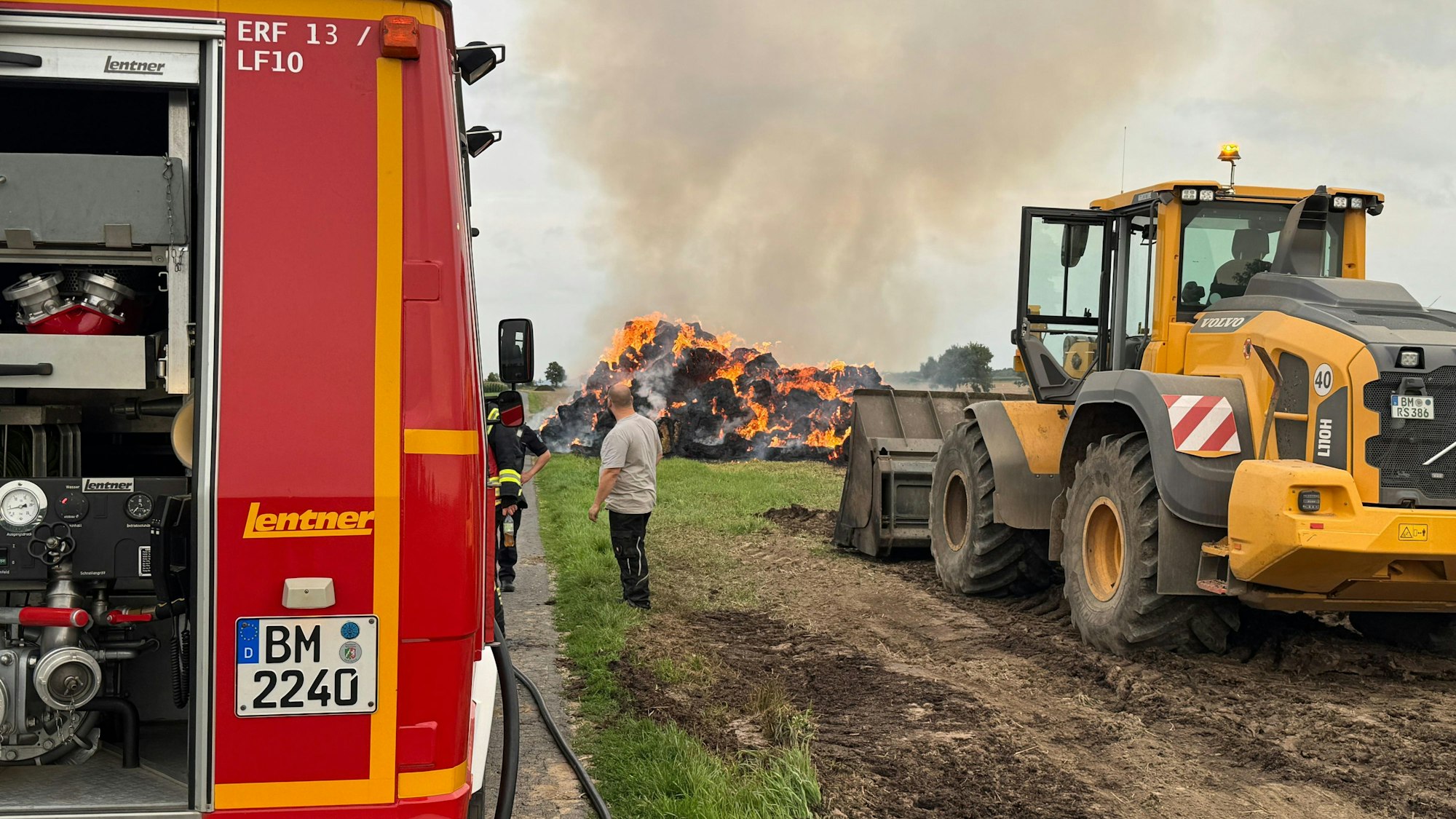 Im Vordergrund ein Feuerwehrfahrzeug und ein Traktor. Im Hintergrund der brennende Haufen Strohballen. In der Mitte des Bildes stehen zwei Personen.