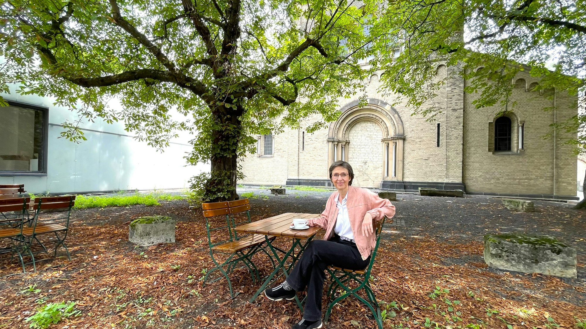 Eine Frau sitzt in einem Innenhof unter einer großen Kastanie an einem Café-Tisch.