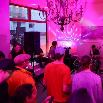 In einem Café spielt eine Band, Menschen stehen im leergeräumten Gastraum und hören zu.