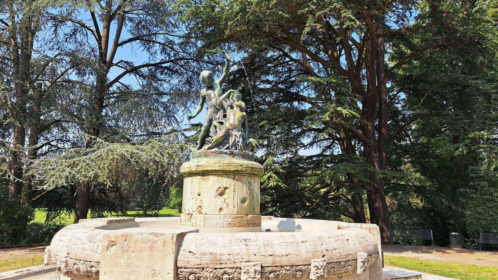 Ein runder steinerner Brunnen steht in einer Grünanlage mit vielen hohen Bäumen.