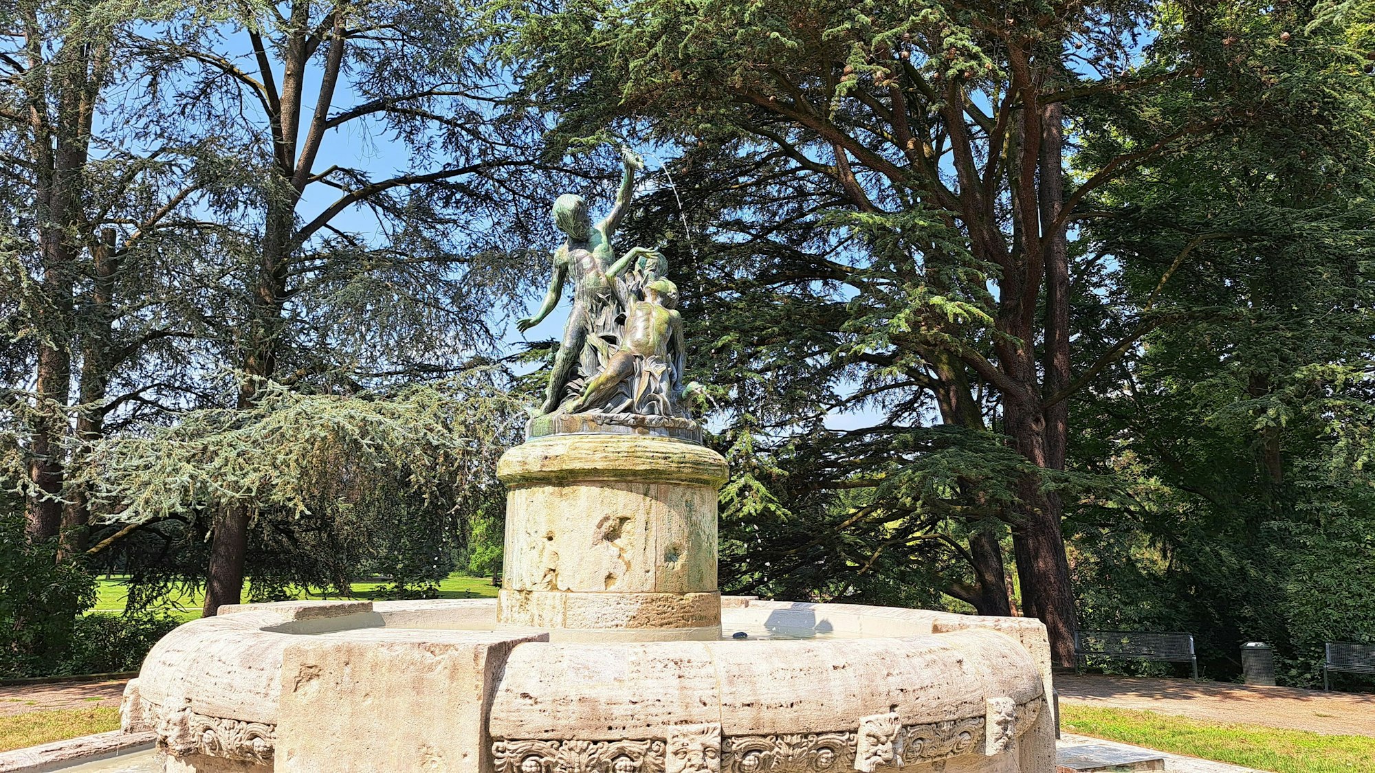 Ein runder steinerner Brunnen steht in einer Grünanlage mit vielen hohen Bäumen.