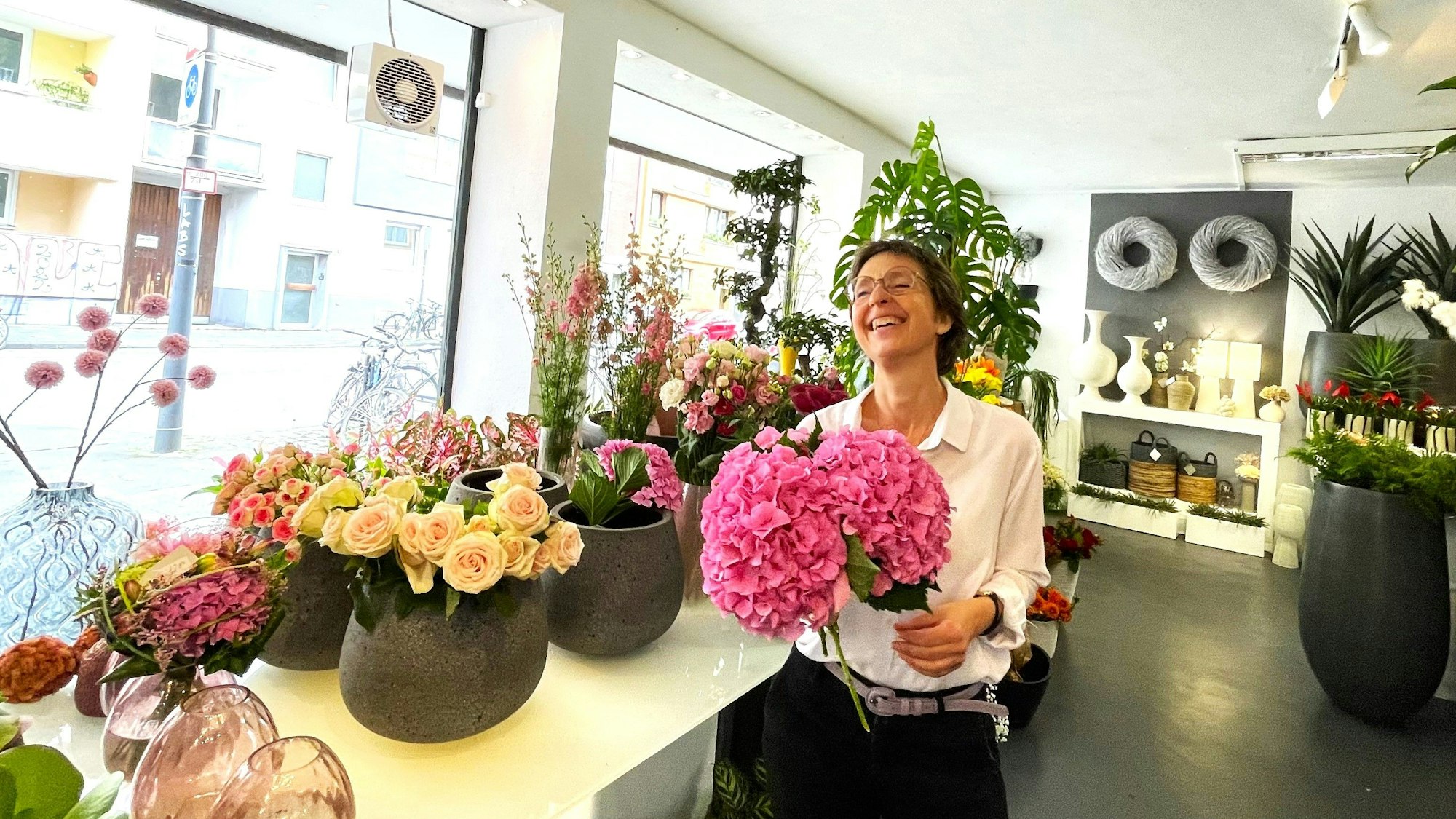 Eine Frau steht in einem Blumengeschäft. Sie hält lachend zwei pinkfarbene Hortensienblüten in der Hand.