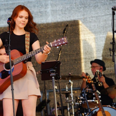 Zu sehen istg eine junge Musikerin mit Gitarre an einem Mikrofon.