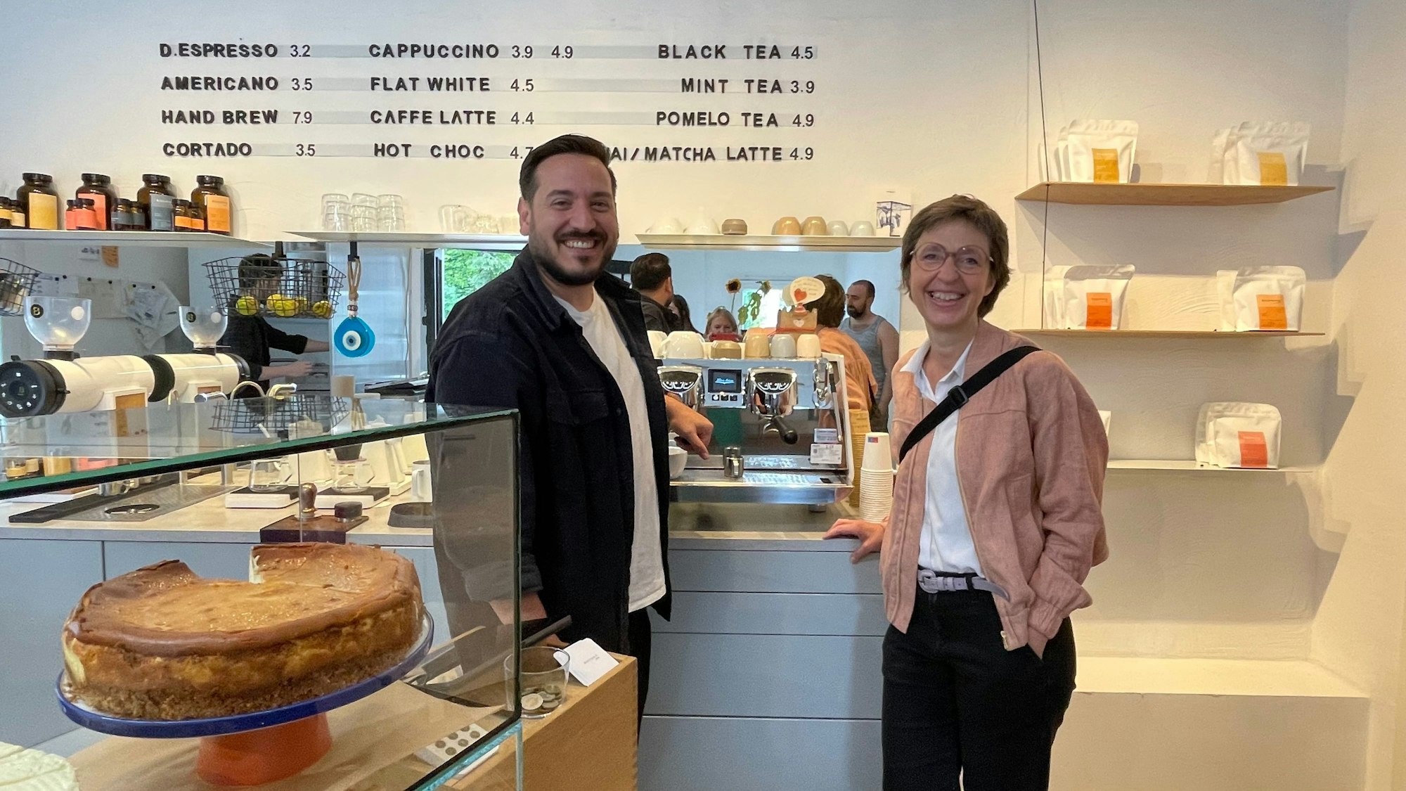 Der Betreiber des Cafés steht neben einer Frau mit Kurzhaarfrisur, die einen lachsfarbenen Blouson trägt.