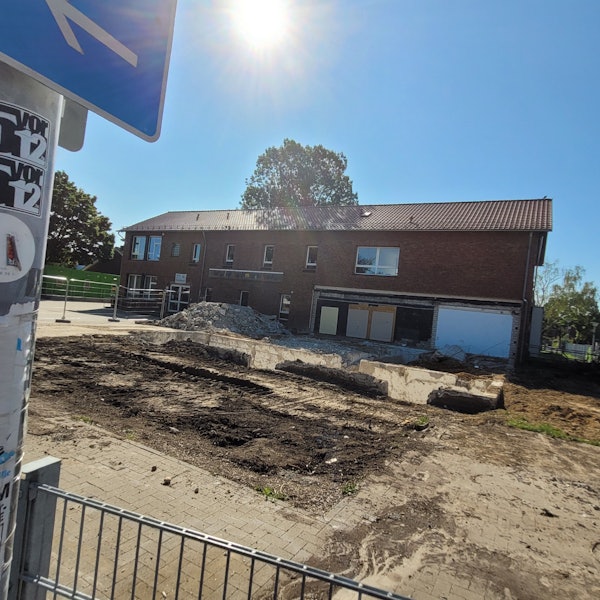 Im Hintergrund ein Gebäude mit Klinkerfassade, davor eine freie Fläche, in der sich noch Mauerreste befinden.